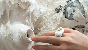 An elegant hand wearing a white color Pear-shape-cut Diamond Square Enamel Ring by www.jingyayi.com.  White textured artistic organic sculptures in the background.