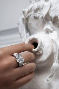 An elegant female hand wearing a scintillating white color Radiant-Cut Moissanite Diamond Ring by www.jingyayi.com.  Well textured artistic organic sculpture in the background.