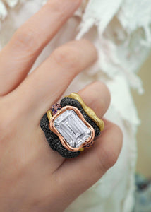 An elegant female hand wearing a crisp sharp 15-carat-size Emerald-Cut Moissanite encrusted on scintillating Black Diamonds, hand made diamond ring by www.jingyayi.com. Well textured pastel color artistic organic sculpture in the background.