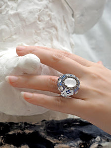 An elegant female hand wearing a white jadeite ring with precious stones handmade by www.jingyayi.com. Well textured white and midnight color artistic organic sculpture in the background.