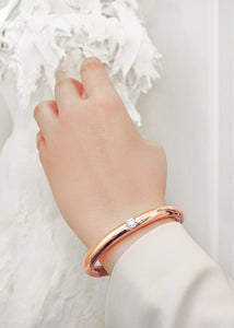 An elegant female wrist wearing a Diamond Bangle in Rose Gold handmade by www.jingyayi.com. Well textured pastel color artistic organic sculpture in the background.