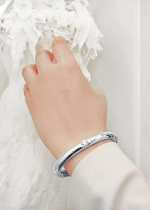 An elegant female wrist wearing a Diamond Bangle in White Gold handmade by www.jingyayi.com. Well textured pastel color artistic organic sculpture in the background.