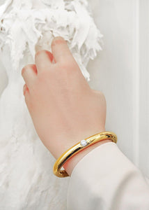 An elegant female wrist wearing a Diamond Bangle in Yellow Gold handmade by www.jingyayi.com. Well textured pastel color artistic organic sculpture in the background.