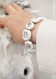 An elegant female hand wearing a scintillating 10-stone Moissanite Enamel Bracelet in bone China white color handmade by www.jingyayi.com. Well textured pastel color artistic organic sculpture in the background.